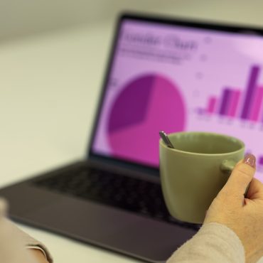A person holding a green cup with a spoon inside while looking at a laptop screen displaying a colorful chart, including a pie chart and bar graph in shades of purple, related to data visualization.