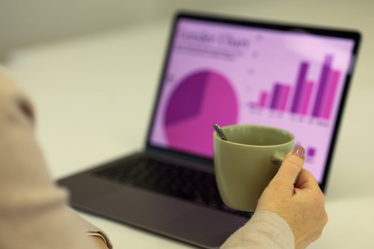 A person holding a green cup with a spoon inside while looking at a laptop screen displaying a colorful chart, including a pie chart and bar graph in shades of purple, related to data visualization.
